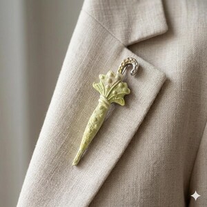 Puede incluir: Broche en forma de paraguas verde claro con un gancho plateado, prendido a una blazer beige. El broche tiene un dise&ntilde;o detallado con un mango texturizado y una parte superior decorativa. La tela de la blazer tiene un sutil patr&oacute;n tejido.