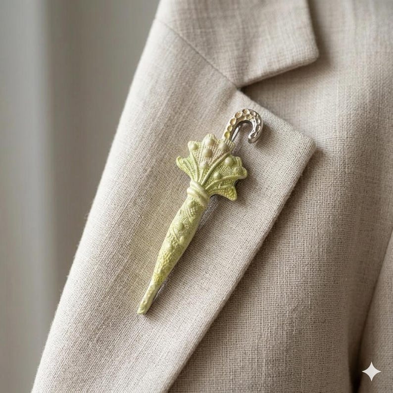 Puede incluir: Broche en forma de paraguas verde claro con un gancho plateado, prendido a una blazer beige. El broche tiene un dise&ntilde;o detallado con un mango texturizado y una parte superior decorativa. La tela de la blazer tiene un sutil patr&oacute;n tejido.