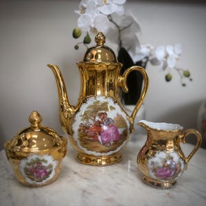 May include: A gold tea set comprising a teapot, sugar bowl, and a creamer. Each item is decorated with a painted scene of figures in a landscape. The set is displayed on a marble surface, with white flowers in the background.