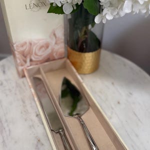 Può includere: Un set di coltello e server per torte argentati in una scatola decorativa. Il set è esposto con fiori bianchi in un vaso di vetro con base dorata. La scatola ha il logo Lenox.