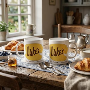 Op de afbeelding: Twee witte keramische mokken met een geel grafisch ontwerp en de bruine tekst Luke's en Stars Hollow, Connecticut op de voorzijde. De mokken staan op een houten tafel naast twee zilveren lepels, croissants en honing.