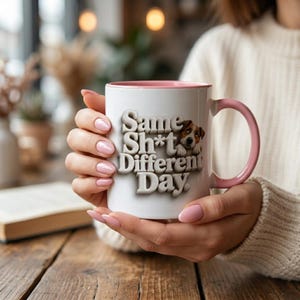 May include: A white ceramic mug with a pink interior and handle, featuring the text "Same Sh*t Different Day" in a 3D font and a small illustration of a brown and white dog peeking out from behind the lettering.