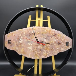 Könnte beinhalten: Eine einzigartige Wanduhr mit einem hellrosa, ovalen Zifferblatt mit eingebetteten floralen Elementen. Das Zifferblatt ist in einem schwarzen kreisförmigen Rahmen eingefasst, der römische Ziffern und schwarze Zeiger mit einem roten Sekundenzeiger anzeigt. Die Uhr wird auf einer goldenen Staffelei präsentiert. Das Zifferblatt ist etwa 25 cm lang.