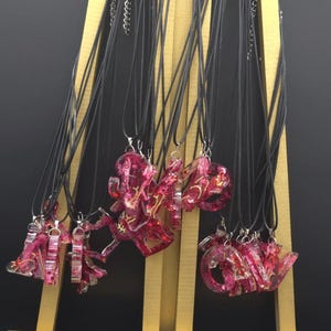 Könnte beinhalten: Eine Sammlung von Buchstabenanhängern aus Harz in Rosa und Gold, präsentiert auf einer kleinen goldenen Staffelei. Jeder Anhänger hängt an einer schwarzen Kordelkette. Die Buchstaben sind transparent mit goldenen Akzenten.