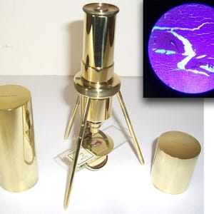 May include: A vintage brass microscope with a tripod base, two cylindrical cases, and a circular inset showing a magnified specimen. The microscope has a long, cylindrical body and a lens at the top. The cases are gold-colored.