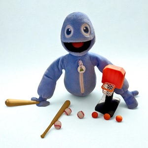 May include: A blue plush toy with a smiling face and a zipper. It is surrounded by miniature sports equipment, including baseball bats, baseballs, a basketball hoop, and orange basketballs. The toy has a small green and yellow logo on the zipper.