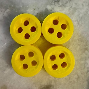 May include: Four bright yellow buttons with six-sided holes arranged in a circular pattern. The buttons are evenly spaced and appear to be made of a solid material. The background is a light gray, textured surface.