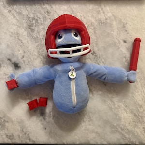 May include: A plush blue figure wearing a red football helmet with a white faceguard. It has a white zipper, a red pencil, and two red squares. The figure has a small pendant with a green and yellow design.