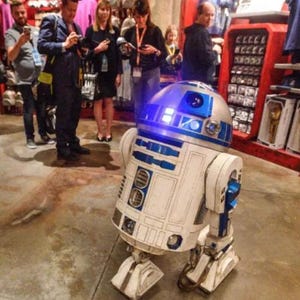 May include: A detailed, life-size R2-D2 droid replica, featuring a silver dome with blue accents and a white body with blue detailing. The droid is on display in a store, with people and merchandise visible in the background.