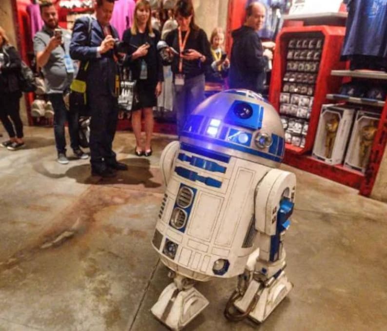 May include: A detailed, life-size R2-D2 droid replica, featuring a silver dome with blue accents and a white body with blue detailing. The droid is on display in a store, with people and merchandise visible in the background.