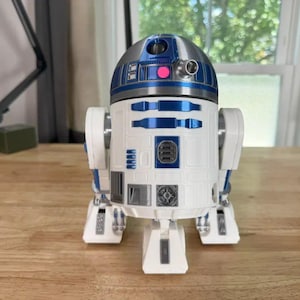 May include: A white, blue, and silver R2-D2 droid figure. The Star Wars character has a domed head with blue and silver accents, a cylindrical body with blue details, and three legs. The figure is on a wooden surface.