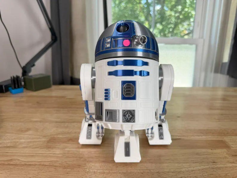 May include: A white, blue, and silver R2-D2 droid figure. The Star Wars character has a domed head with blue and silver accents, a cylindrical body with blue details, and three legs. The figure is on a wooden surface.
