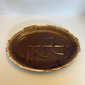 May include: Oval brown ceramic serving platter with a leaf design in the center. The platter has a dark brown interior and a lighter brown and white rim. The leaf design is raised and adds texture to the dish.