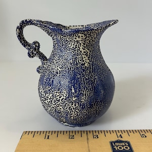 Peut inclure: Une petite cruche en céramique bleue et blanche tachetée, avec une anse incurvée et un bord évasé. La cruche a une surface texturée et un corps bulbeux. Une règle en bois est placée à côté.