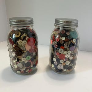 May include: Two clear glass mason jars with silver metal lids, each filled with a large collection of assorted sewing buttons in various colors, shapes, and sizes, including red, blue, green, brown, black, white, and metallic tones.