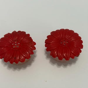 May include: A pair of round, bright red salt and pepper shakers shaped like stylized daisies. Each shaker features a textured floral petal design on the top surface with a small cluster of holes in the center for dispensing seasoning.
