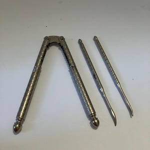 May include: A silver-toned metal nutcracker with a textured, cross-hatched pattern on the handles and two matching slender metal picks with pointed, curved ends. The nutcracker features a hinged design with a serrated inner grip area.