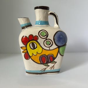 May include: A ceramic pitcher with a whimsical rooster design. The pitcher features a cream-colored base with a colorful rooster illustration in yellow, red, and purple. It has a spout, handle, and is accented with blue and brown stripes. A unique decorative item.