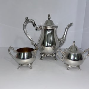 May include: A silver-toned tea set consisting of a teapot, a creamer, and a lidded sugar bowl. Each piece features ornate, curved handles and decorative feet. The teapot has a tall, pointed lid, while the sugar bowl has a matching lid.