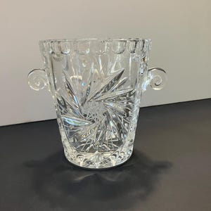 May include: A clear, cut-glass ice bucket with a starburst design on the front. The bucket has two decorative handles and a scalloped rim. The glass reflects light, creating a sparkling effect. This vintage piece is perfect for entertaining.