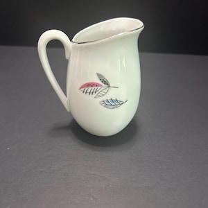 May include: A white porcelain creamer pitcher featuring a curved handle and a silver-toned rim. The body is decorated with a small graphic pattern of two stylized leaves, one in pink and one in blue, each accented with fine black line details.