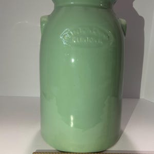 May include: A light green ceramic jar with a glossy finish. The jar has a cylindrical shape with a slightly flared top and two small handles. The text "ANTONIO CLINTON, IL" is embossed on the front. A wooden ruler is at the bottom.