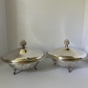 May include: Two silver-toned serving bowls with matching lids, each featuring a polished finish, a beaded rim detail, and a decorative shell-shaped finial on top. The bowls are supported by three ornate, curved feet, providing a raised base.