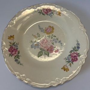 May include: A round, off-white ceramic plate with a scalloped edge and a glossy finish. The center and rim feature floral arrangements with pink roses, yellow tulips, and small blue flowers, all accented with green leaves and stems.