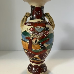 May include: A ceramic vase with a cream-colored base featuring two handles and hand-painted designs in red, green, blue, and black. The artwork depicts a figure in traditional attire surrounded by abstract patterns, floral motifs, and white dots.