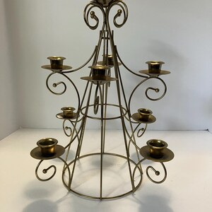 May include: A gold-toned metal candelabra featuring a tiered, conical frame with nine candle holders. The structure is composed of thin, curved wire arms that spiral outward, supporting circular drip plates at varying heights in a symmetrical arrangement.