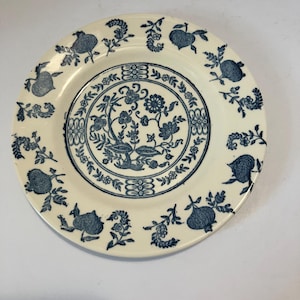 May include: A round ceramic plate with a cream-colored base and a blue transferware pattern. The center features a floral motif surrounded by a geometric border, while the rim is decorated with a repeating pattern of pomegranates and leaves.