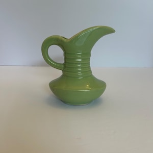 May include: A small, light green ceramic pitcher featuring a glossy finish, a curved handle, and a wide, flared spout. The neck of the pitcher is decorated with three horizontal, raised ridges that wrap around the circumference of the vessel.