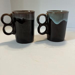 May include: Two ceramic mugs with dark brown bodies and unique double-loop handles. The left mug features a greyish-brown drip glaze at the rim, while the right mug has a distinct light blue drip glaze at the rim, both with a glossy finish.