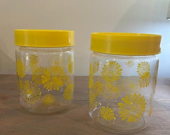 Pode incluir: Dois frascos de vidro transparente com tampas amarelas. Os frascos são decorados com um padrão floral amarelo. Os frascos estão sobre uma superfície de madeira. Os frascos são provavelmente para armazenamento na cozinha.