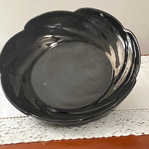 May include: A glossy black ceramic bowl with a scalloped, petal-like rim. The bowl features a smooth, reflective surface and a deep, rounded basin, showcasing a dark monochromatic finish with subtle light reflections across its curved edges.