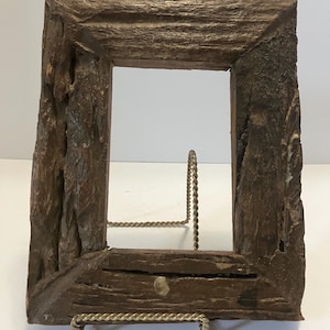 May include: A rectangular picture frame crafted from dark brown, textured wood with a rough, bark-like surface. The frame features mitered corners and is supported by a thin, gold-toned metal wire stand that holds it in an upright position.