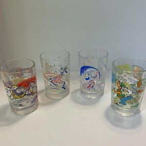 May include: Four clear glass tumblers featuring colorful Disney character graphics. The glasses display Buzz Lightyear, Goofy, Mickey Mouse, and Goofy again, with various printed text and illustrations including the Walt Disney World logo.