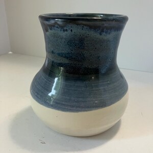May include: A ceramic vase with a rounded base and a flared rim. The bottom section is a solid cream color, while the upper portion features a deep blue glaze that transitions into a darker, speckled navy tone near the top opening of the vessel.