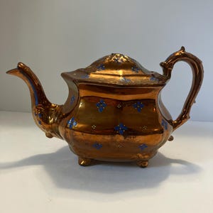 May include: A metallic copper-colored teapot featuring a glossy finish and a repeating pattern of small blue four-petaled flowers with yellow centers. The teapot has a curved handle, a pointed spout, a lid, and stands on four small feet.
