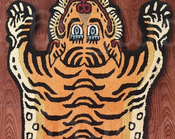 May include: A rug shaped like a tiger, featuring orange and black stripes. The rug has a detailed face with blue eyes and a cream-colored border. The rug is placed on a wooden floor.