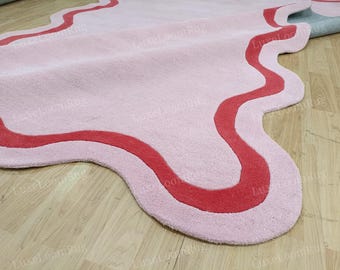 May include: A pink rug with a wavy, irregular shape and a red border. The rug is made of a soft, plush material and is placed on a light-colored wooden floor. The design is modern and abstract.