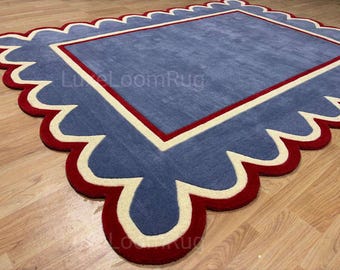 May include: A rectangular rug with a scalloped edge design. The rug features a blue center, a red border, and a cream-colored accent. The rug is placed on a wooden floor.