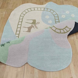 May include: A whimsical, abstract-shaped rug in pastel colors. The rug features a light blue, green, and lavender color scheme with a beige section that has a ladder and animal design. The rug is on a wooden floor.