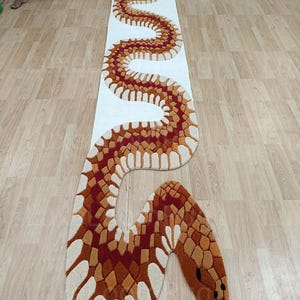 Könnte beinhalten: Ein langer, schlangenförmiger Teppich mit weißem Hintergrund und detailliertem Muster. Der Körper der Schlange weist ein sich wiederholendes Design aus roten, orangefarbenen und beigefarbenen Formen auf. Der Schlangenkopf hat eine rote Zunge und schwarze Augen.