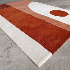 May include: A rectangular rug with a geometric design. The rug features a large section of rust-colored material, a white border, and a white semi-circle with a rust-colored circle. The rug is made of a soft, plush material.