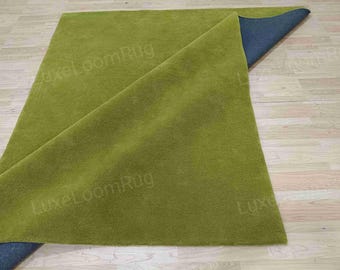 Puede incluir: Una alfombra rectangular verde con una esquina doblada, que revela un reverso azul oscuro. La alfombra está sobre un suelo de madera clara. La textura de la alfombra parece suave y afelpada, adecuada para la decoración del hogar.