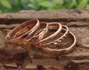 Peut inclure: Un ensemble de cinq bagues en or rose aux textures variées. Les bagues sont posées sur une surface brune texturée, avec un feuillage vert en arrière-plan. Les bagues présentent des motifs martelés, texturés et lisses.