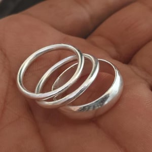 May include: Three silver rings of different widths are held in an open palm. The rings are smooth and shiny, reflecting light. The rings appear to be made of sterling silver and are likely worn as jewellery.