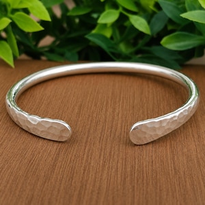 May include: A silver cuff bracelet with a polished finish and a hammered texture. The bracelet features an open design with rounded ends, showcasing a reflective metallic surface that catches the light across its curved, textured exterior.