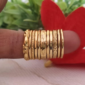May include: A set of ten gold-toned rings featuring various textures and patterns, including smooth bands, floral engravings, and geometric designs, arranged side-by-side to display the distinct surface details of each individual metal ring.
